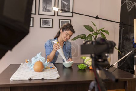 Photo for Vlogger filming video about introducing soft food to baby nutrition and preparation of fresh fruit and vegetable porridges using stick mixer as part of online prenatal classes course - Royalty Free Image