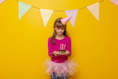 Photo for Beautiful little girl wearing birthday hat, holding a birthday cake with lit candles on it, making a wish and blowing candles, isolated on yellow colored background - Royalty Free Image