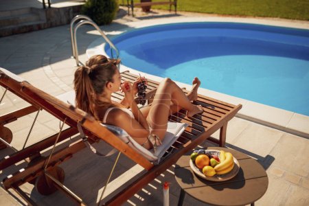 Téléchargez les photos : Femme allongée sur un transat au bord de la piscine, mangeant du raisin, bronzant et se relaxant pendant les vacances d'été - en image libre de droit