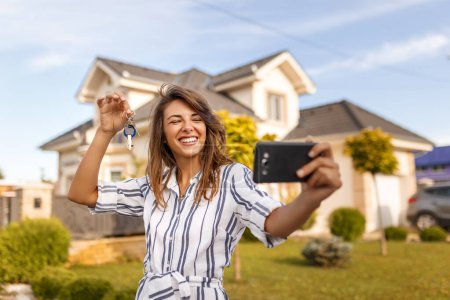 Téléchargez les photos : Belle jeune femme debout devant sa nouvelle maison, prenant un selfie en utilisant un téléphone intelligent avec les clés de sa propriété nouvellement achetée - en image libre de droit