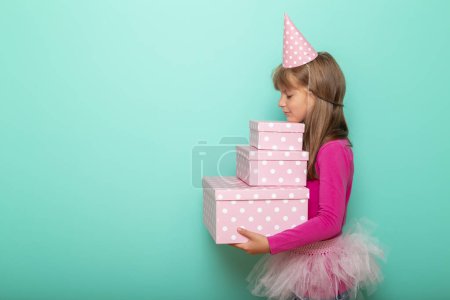 Photo for Beautiful little girl wearing party hat, celebrating her birthday and holding a bunch of birthday presents, isolated on mint colored background - Royalty Free Image