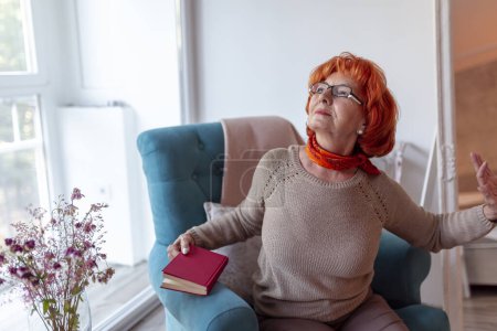 Téléchargez les photos : Femme âgée assise dans un fauteuil, étirant et bâillant en lisant un livre - en image libre de droit