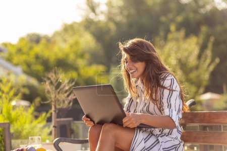 Téléchargez les photos : Belle jeune femme se détendre sur la terrasse de la cour, avoir un appel vidéo à l'aide d'un ordinateur portable et profiter de la journée ensoleillée d'été en plein air - en image libre de droit