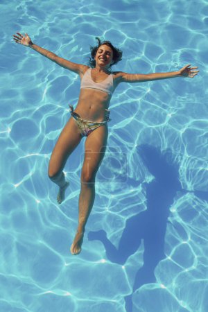 Téléchargez les photos : Vue en angle élevé de belle jeune femme portant un maillot de bain flottant sur l'eau dans la piscine se détendre pendant les vacances d'été - en image libre de droit
