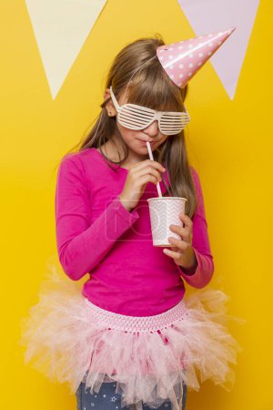 Foto de Hermosa niña con gafas de fiesta y sombrero de cumpleaños, beber jugo con una paja, aislado en el fondo de color amarillo con banderas de fiesta - Imagen libre de derechos