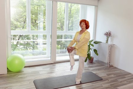 Téléchargez les photos : Femme âgée active faisant de l'entraînement de yoga à la maison, debout près du tapis de yoga, pratiquant l'équilibre concept de mode de vie sain - en image libre de droit