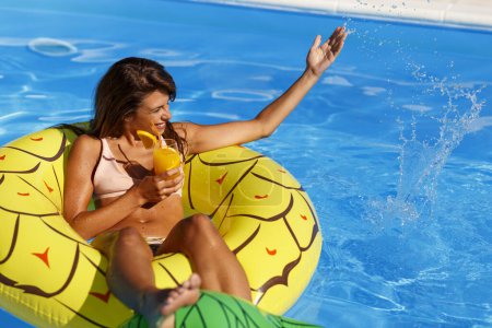 Téléchargez les photos : Attrayant jeune femme en maillot de bain boire cocktail tout en flottant sur le radeau de la piscine, profiter de la journée ensoleillée d'été à la piscine - en image libre de droit