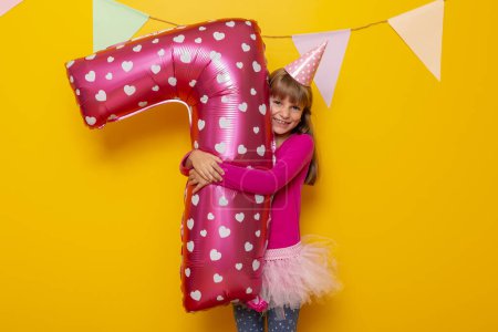 Photo for Portrait of a beautiful little girl having fun while celebrating birthday, holding a giant pink balloon shaped as number seven, isolated on yellow colored background - Royalty Free Image