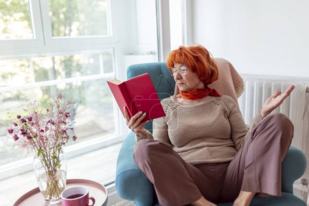 Foto de Hermosa mujer mayor sentada en un sillón junto a la ventana, leyendo un libro y disfrutando del tiempo libre en casa - Imagen libre de derechos