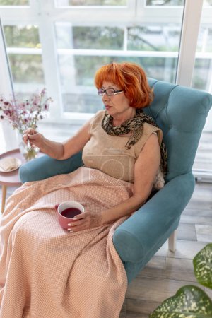 Foto de Mujer anciana enferma sentada en un sillón junto a la ventana cubierta con manta, con gripe y fiebre, midiendo la temperatura usando termómetro y tomando una taza de té caliente - Imagen libre de derechos