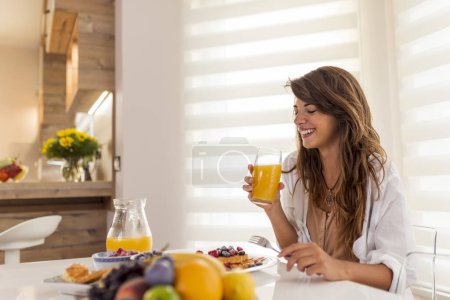 Téléchargez les photos : Jeune femme assise à la table de la salle à manger et souriante, buvant du jus d'orange frais pour le petit déjeuner - en image libre de droit