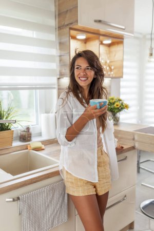 Photo for Beautiful young woman standing in the kitchen, drinking morning coffee and enjoying leisure time at home - Royalty Free Image