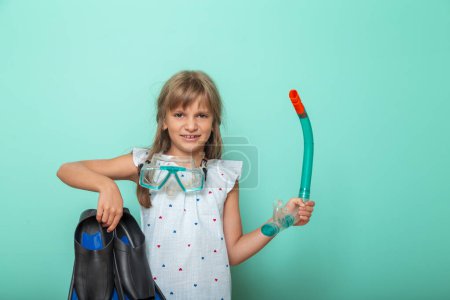 Téléchargez les photos : Petite fille excitée se préparant pour les vacances d'été à la plage, tenant une paire de palmes pleines pieds et masque de plongée, isolé sur fond de couleur menthe - en image libre de droit