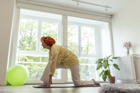 Téléchargez les photos : Femme âgée pratiquant le yoga à la maison ; mode de vie sain et actif des personnes âgées concept - en image libre de droit