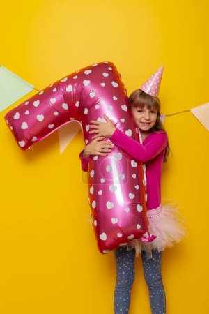 Foto de Retrato de una hermosa niña divirtiéndose mientras celebra su cumpleaños, sosteniendo un globo rosa gigante en forma de número siete, aislado sobre fondo de color amarillo - Imagen libre de derechos