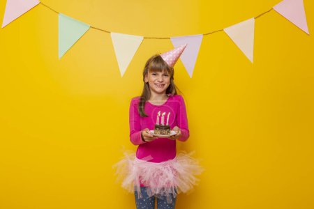 Téléchargez les photos : Belle petite fille portant un chapeau d'anniversaire, tenant un gâteau d'anniversaire avec des bougies allumées dessus, faisant un souhait et soufflant des bougies, isolé sur fond jaune - en image libre de droit