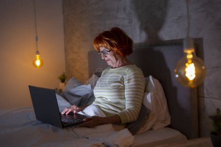 Photo for Beautiful senior woman lying in bed at night, having a video call on a laptop computer with family members - Royalty Free Image