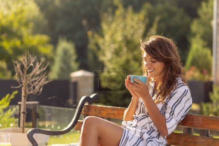 Téléchargez les photos : Belle jeune femme se détendre sur la terrasse dans la cour de sa maison, boire du café le matin et profiter de la journée ensoleillée d'été en plein air - en image libre de droit