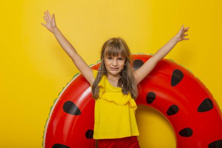 Photo for Little girl standing in front of an inflatable watermelon swim ring, getting ready for beach summer vacation, isolated on yellow colored background - Royalty Free Image