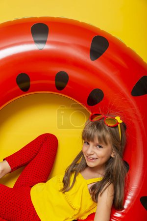 Téléchargez les photos : Petite fille assise dans un anneau de natation gonflable de pastèque, se préparant pour les vacances d'été à la plage, isolée sur fond jaune - en image libre de droit