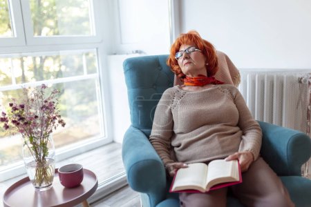 Photo for Senior woman sleeping in an armchair with a book on her lap, having an afternoon nap and relaxing at home - Royalty Free Image