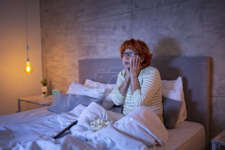Foto de Hermosa mujer mayor con pijamas sentada en la cama, comiendo palomitas de maíz y viendo películas de terror en la televisión, asustada y tensa - Imagen libre de derechos