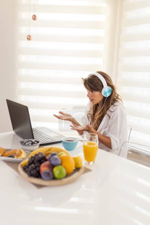 Téléchargez les photos : Belle jeune femme assise à la table de salle à manger, portant un casque et ayant un appel vidéo à l'aide d'un ordinateur portable tout en prenant le petit déjeuner à la maison - en image libre de droit
