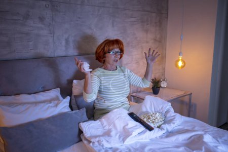 Téléchargez les photos : Femme âgée en pyjama assise sur le lit, regardant un film dramatique à la télévision, surprise - en image libre de droit