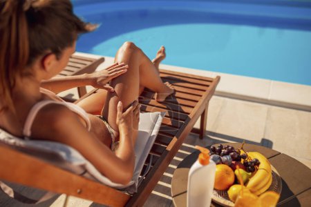 Téléchargez les photos : Belle jeune femme bronzant au bord de la piscine pendant les vacances d'été, appliquant de la crème solaire sur sa peau comme protection solaire contre les rayons UVA et UVB - en image libre de droit