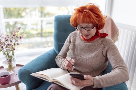 Photo for Beautiful senior woman relaxing at home, sitting in an armchair, drinking tea, using a smart phone and writing in a planner - Royalty Free Image