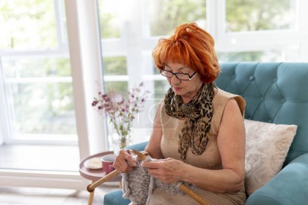 Foto de Mujer mayor disfrutando de su tiempo libre en casa, sentada en un sillón, tejiendo una bufanda - Imagen libre de derechos