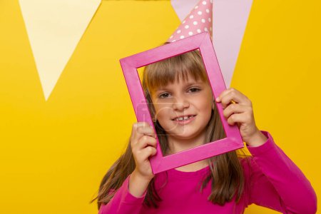 Photo for Beautiful little girl having fun celebrating her birthday, holding a pink photo frame and posing for a photo, isolated on yellow colored background - Royalty Free Image