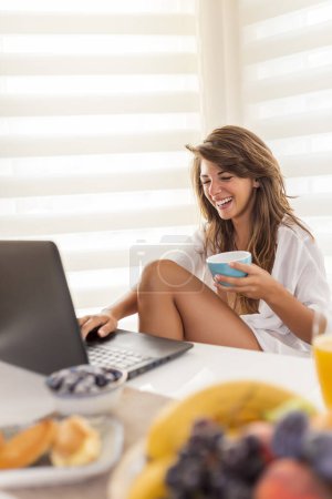 Photo for Beautiful young woman sitting at dinning room table, using laptop computer while having breakfast at home in the morning - Royalty Free Image