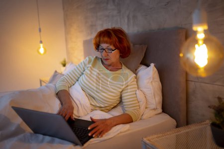 Téléchargez les photos : Portrait d'une femme âgée portant un pyjama couchée la nuit, travaillant tard à l'aide d'un ordinateur portable - en image libre de droit