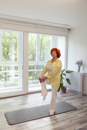 Foto de Mujer mayor activa haciendo entrenamiento de yoga en casa, de pie junto a la esterilla de yoga, practicando el equilibrio - concepto de estilo de vida saludable - Imagen libre de derechos