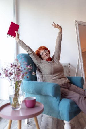 Photo for Senior woman sitting in an armchair, stretching and yawning while reading a book - Royalty Free Image