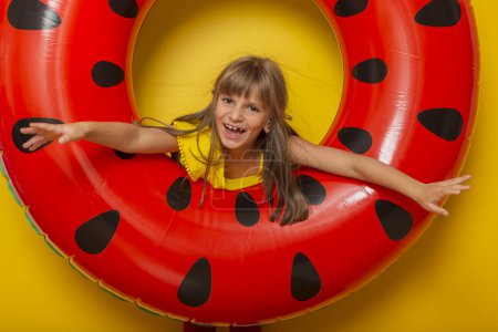 Téléchargez les photos : Petite fille jouant avec un anneau gonflable de natation de pastèque, se préparant pour des vacances d'été à la plage, isolé sur fond jaune - en image libre de droit