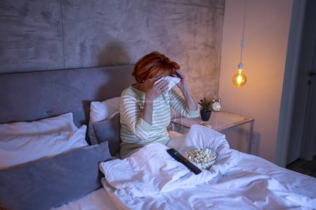 Téléchargez les photos : Femme âgée portant un pyjama assis sur le lit, regardant un film dramatique à la télévision, triste, pleurant et essuyant des larmes avec un mouchoir - en image libre de droit