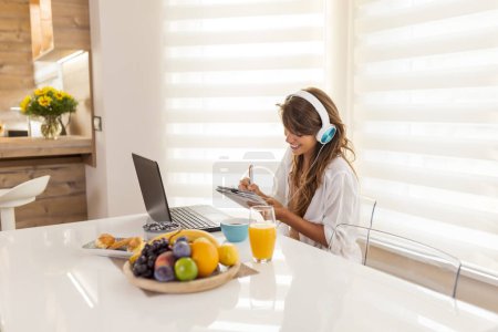 Photo for Woman taking an online educational course, wearing headset and writing in a planner while listening to a lesson - Royalty Free Image