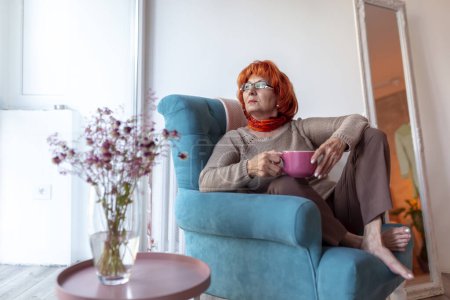 Téléchargez les photos : Belle femme âgée assise dans un fauteuil près de la fenêtre, buvant du thé et regardant par la fenêtre, se relaxant à la maison - en image libre de droit