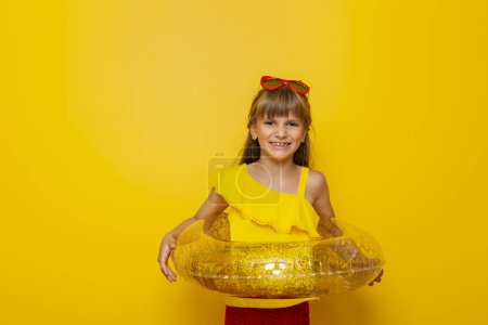 Téléchargez les photos : Petite fille tenant un anneau de natation gonflable, se préparant pour les vacances d'été à la plage, isolée sur fond jaune - en image libre de droit