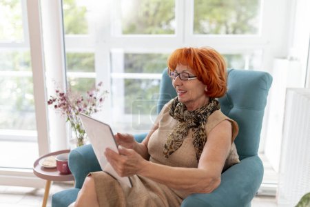 Photo for Elderly woman sitting in an armchair, relaxing at home and using a tablet computer; senior woman surfing the Net on tablet computer - Royalty Free Image