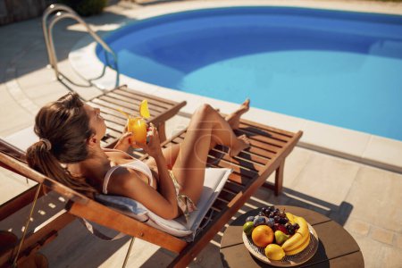 Foto de Hermosa mujer tumbada en la cama de sol junto a la piscina, bebiendo cócteles y tomando el sol durante las vacaciones de verano - Imagen libre de derechos