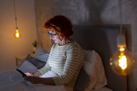Téléchargez les photos : Femme âgée en pyjama assise sur le lit, tapant un message texte sur un téléphone intelligent - en image libre de droit