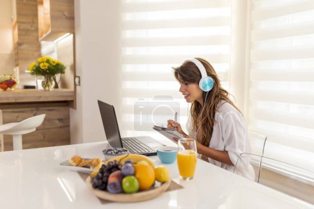 Téléchargez les photos : Femme travaillant à distance de la maison portant un casque et prenant des notes tout en ayant une réunion en ligne - en image libre de droit