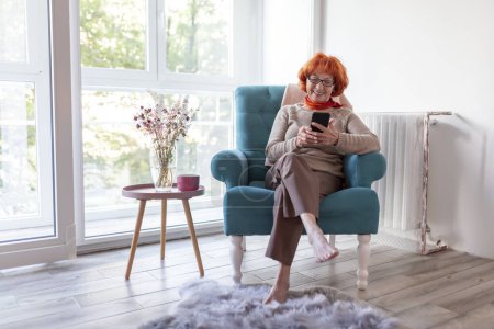 Téléchargez les photos : Femme âgée profitant de son temps libre à la maison, assise dans un fauteuil et ayant un appel vidéo à l'aide d'un téléphone intelligent - en image libre de droit