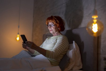 Téléchargez les photos : Femme âgée portant un pyjama assis sur le lit, tapant un message texte à l'aide d'un téléphone intelligent - en image libre de droit