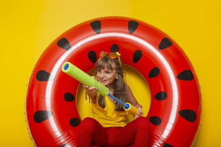 Téléchargez les photos : Petite fille jouant avec le jouet de pistolet à eau dans un anneau gonflable de natation de pastèque, se préparant pour les vacances d'été de plage, isolé sur fond jaune - en image libre de droit