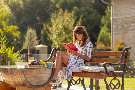 Schöne junge Frau genießt sonnigen Sommermorgen im Freien, trinkt Kaffee und liest ein Buch auf der Hinterhofterrasse