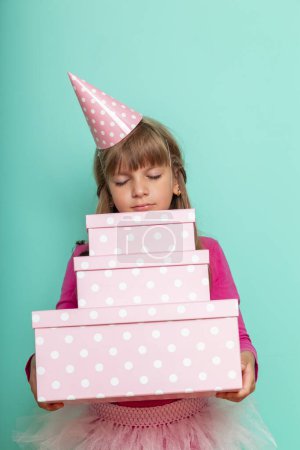 Téléchargez les photos : Belle petite fille portant un chapeau de fête, célébrant son anniversaire et tenant un tas de cadeaux d'anniversaire, isolé sur fond de couleur menthe - en image libre de droit